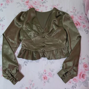 Olivgrön satinblus med volang - Supersnygg olivgrön blus i glansigt satinmaterial med v-ringning, långa puffärmar och knappar framtill. Croppad modell med volangkant nedtill och breda manschetter med knappar. Perfekt för dig som vill sticka ut med en trendig och elegant look.