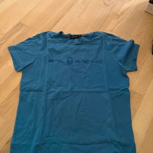 Blå Sail Racing t-shirt i bomull - Blå t-shirt från Sail Racing med tryckt logga framtill. Klassisk rund halsringning och korta ärmar. Tillverkad i mjuk bomull, perfekt för en avslappnad stil. Snygg och enkel design som passar till jeans eller shorts.