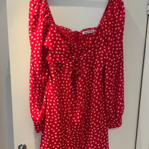 Röd blommig Mykonos klänning från Adoore - Supersöt röd kortklänning från Adoore med vita blommor, snörning framtill och långa ärmar. Klänningen har en smickrande v-ringning och dragkedja i sidan. Perfekt för sommarens alla tillfällen!