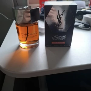 YSL L'Homme Eau de Parfum 100ml - YSL L'Homme Eau de Parfum i 100 ml flaska. Parfymen kommer i en stilren glasflaska med bärnstensfärgad vätska och en stor, sexkantig silverfärgad metallkork. Medföljer originalkartong i svart och silver med YSL-logga.
