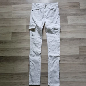 Vita cargopants från UNIQ MINI - Snygga vita cargopants från UNIQ MINI i storlek 170. Byxorna har flera praktiska fickor på sidorna och klassisk cargostil. De är raka i modellen och har bälteshällor samt dragkedja och knapp framtill. Perfekta för en clean och trendig look.