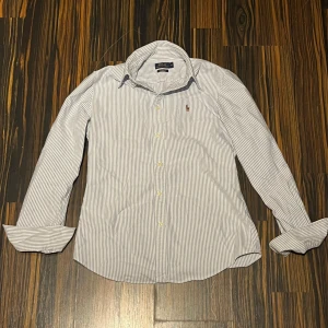 Randig skjorta från Polo Ralph Lauren - Snygg skjorta från Polo Ralph Lauren i vitt och blått med tunna ränder. Klassisk krage, långärmad och med broderad logga på bröstet. Custom fit-modell som sitter riktigt bra. Perfekt nu under sommarn, säljer för att den är för liten, annars är den riktigt skön och snygg. Inte fast pris. Gärna snabb affär