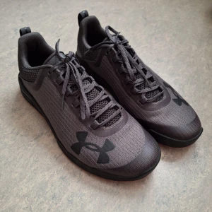 Svarta Under Armour sneakers - Snygga svarta sneakers från Under Armour med diskret logga på sidan och tå. Ovandel i mesh och syntet för bra andningsförmåga, samt röda detaljer på sulan som ger en sportig vibe. Perfekta för träning eller chill vardagsstil.