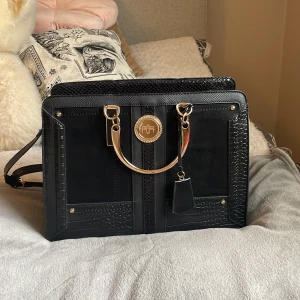 Svart handväska från River Island - Stilren svart handväska från River Island med guldfärgade handtag och detaljer. Väskan har strukturmönstrade partier, flera fack och dragkedja upptill. Snygg mix av slätt och präglat syntetmaterial, perfekt för dig som gillar statement-accessoarer. Knappt använd och endast lite slitage i ett av hörnen! Bor du i närheten kan jag frakta väskan till dig 💕