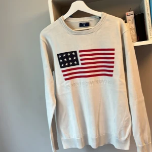 GANT Vit stickad tröja med USA-flagga - Vit stickad tröja från GANT i storlek M med broderad amerikansk flagga på bröstet. Tröjan har rund halsringning, ribbade muddar och är långärmad. Perfekt för dig som gillar preppy stil och vill sticka ut med en klassisk detalj.