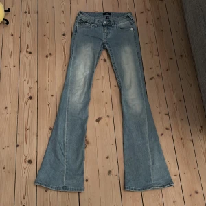 True Religion Joey Low Rise Flare Jeans - Snygga ljusblå jeans från True Religion, modell Joey Low Rise Flare. De har låg midja, utsvängda ben och klassiska detaljer som markerade sömmar och fickor med lock och knappar bak. Perfekta för dig som gillar retrovibbar och statement-jeans. Knappt använda