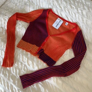 Färgglad randig kofta från Collusion - Säljer en croppad kofta från Collusion i röd, orange och mörkblå med gula detaljer. Tröjan är ribbstickad, har v-ringning, knappar framtill och långa ärmar.