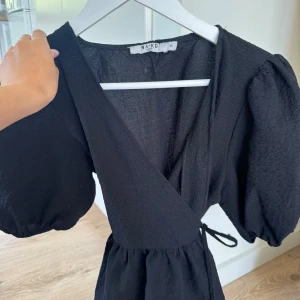 Svart omlotttopp från NA-KD - Supersnygg svart omlotttopp från NA-KD med puffiga korta ärmar och knyt i midjan.  har en lätt strukturerad yta och v-ringad hals. Perfekt för dig som gillar en trendig och stilren look.