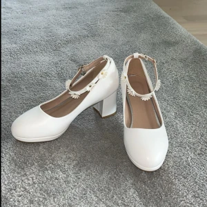 Vita klackskor med blommiga detaljer - Söta vita klackskor med rund tå och blockklack. Skorna har en rem runt ankeln dekorerad med små vita blommor och gulddetaljer. Perfekta för dig som vill ha en gullig och stilren look. Tillverkade i syntetmaterial med mjuk innersula. Aldrig använda. 