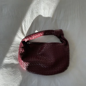 flätad axelväska  - Säljer en fin vinröd axelväska i flätat skinn med rundad form och bred axelrem med knutdetalj. Väskan har dragkedja upptill och en riktigt snygg, trendig look som passar till det mesta. Perfekt för dig som gillar stilrena accessoarer med det lilla extra. 💌