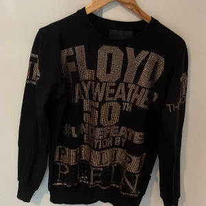 Svart Philipp Plein x Mayweather tröja - Svart långärmad tröja från Philipp Plein med stora glittriga gulddetaljer och texten 'Floyd Mayweather 50th Undefeated Edition'. Tröjan har crewneck, ribbade muddar och är dekorerad med strass på både fram- och baksida samt ärmar.