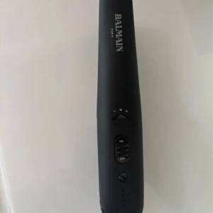 Balmain Cordless Straightener - Snygg och smidig sladdlös plattång från Balmain i matt svart design med silvriga plattor. Perfekt för att styla håret on-the-go, med flera värmelägen och enkel on/off-knapp. Lyxig känsla och passar i väskan. Det är i bra skick.