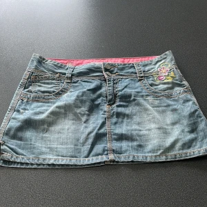 Jeanskjol med broderi från Rip Curl - Snygg ljusblå jeanskjol från Rip Curl med broderade blommor i rosa, lila och gult på både fram- och baksida. Kjolen har klassiska jeansfickor, synliga sömmar och små slitsar i sidorna. Perfekt för dig som gillar en chill och färgglad stil.