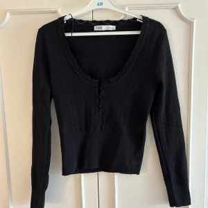 Svart stickad topp med knappar från Zara - Supersnygg svart stickad topp från Zara med djup rundad ringning och dekorativa knappar framtill. Långärmad och figurnära passform, perfekt till jeans eller kjol. Mjukt material som känns skönt mot huden. Använd en gång endast, liten M