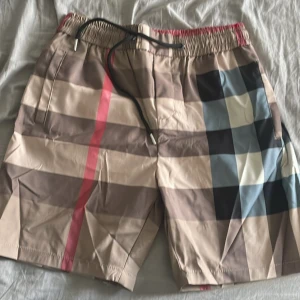 Rutigta badshorts med snörning - Snygga badshorts med klassiskt rutigt mönster i beige, brunt, svart, blått och vitt samt en röd rand. Shortsen har elastisk midja med svart snörning och sidofickor. Perfekta för stranden eller poolen, riktigt trendig vibe.