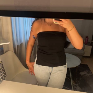 Svart bandeau tubtopp - Säljer en svart bandeau tubtopp med enkel och stilren design. Toppen är axelbandslös och har en liten slits på sidan som ger en snygg detalj. Perfekt att matcha med jeans eller kjol för en clean look.