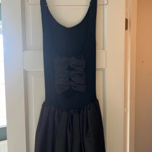 Svart kort klänning från Hunza - Supersöt svart kort klänning från Hunza med smockad överdel och voluminös kjol. Baksidan har tre stora rosetter som ger en unik look. Klänningen är ärmlös och gjord i ribbat tyg som en baddräkt, perfekt för dig som vill sticka ut.