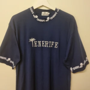 Vintage T-shirt 90-tal - Mörkblå t-shirt från Namaste med vit brodyr 'Tenerife' och palm på bröstet. Kragen och ärmsluten har vit text och motiv. Skön passform och perfekt för sommarens äventyr. Klassisk rund hals och korta ärmar.