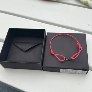 Valentino Garavani rosa armband - Snyggt armband från Valentino Garavani med starkt rosa snöre och ikonisk V-logga i silverfärgad metall. Justerbar passform med knutar och levereras i original ask. Perfekt accessoar för att lyfta din stil.