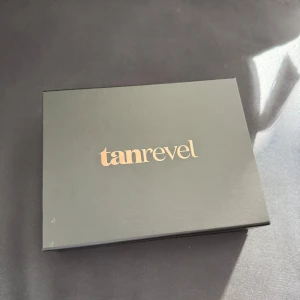 Tanrevel Tanning Kit - Tanrevel Tanning Kit med en svart, matt apparat för brun-utan-sol, tillhörande Tanning Liquid och USB-laddningskabel. Snygg och smidig design, perfekt för att få en solkysst look året runt. Kommer i elegant svart förpackning med guldtext. Endast provad