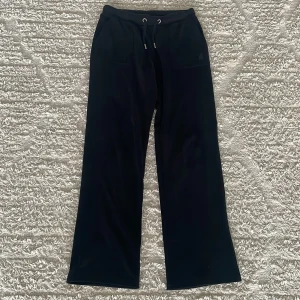 Svarta velourbyxor Juicy Couture - Svarta mjukisbyxor från Juicy Couture i mjuk velour med snörning i midjan och broderad logga framtill. Byxorna har raka ben och fickor både fram och bak med knappdetaljer. Perfekta för en chill och trendig look.