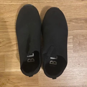 Svarta slip-on sneakers i mesh - Svarta sneakers med slip-on design och rund tå. Ovandelen är i stickad mesh som ger en sportig och clean look. Skorna har en platt sula i svart gummi och är perfekta för en avslappnad streetstyle.