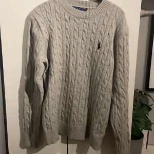 Grå kabelstickad tröja Polo Ralph Lauren - Kabelstickad grå tröja från Polo Ralph Lauren med klassisk rund hals och broderad svart logga på bröstet. Tröjan är nästan aldrig använd. Långärmad och ribbade muddar. Perfekt för dig som gillar stilren och tidlös design. Skriv vid minsta fundering!