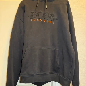 Svart hoodie från Hugo Boss - Svart hoodie från Hugo Boss med broderad logga i svart och orange framtill. Klassisk känguruficka, huva med snörning och mjukt material som känns skönt mot huden. Perfekt för en chill och stilren look. (Dock inte äkta) 