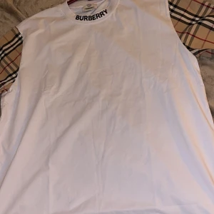 Vit Burberry t-shirt med rutiga ärmar - Snygg vit t-shirt från Burberry med klassiska rutiga ärmar i beige, svart och rött. Rund halsringning med Burberry-logga. Perfekt för dig som vill sticka ut med en ikonisk design och streetstyle-vibe. Finns i alla storlekar 