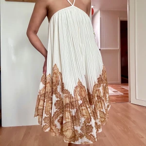 Pleated halter summer dress - Superfin plisserad kortklänning från H&M i vitt med beige paisleymönster nertill. Klänningen har tunna axelband och luftig passform, perfekt för varma dagar. Materialet känns lätt och flowy, och mönstret ger en bohemisk vibe.