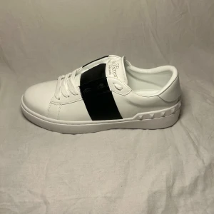 Valentino Garavani Open sneakers vit/svart - Snygga Valentino Garavani Open sneakers i vitt skinn med bred svart läderdetalj över mitten. Klassisk rund tå, vita snören och coola nitar längs hälen. Guldlogga på plösen och innersulan. Perfekta för dig som gillar lyxig streetstyle.