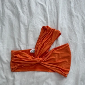 Orange oneshoulder topp från Dalilis - Snygg orange oneshoulder topp från Dalilis med knut framtill. Gjord i mjuk bomull, perfekt för att sticka ut med en cool och trendig look. Toppen har en ärmlös sida och en axelbandslös sida för en unik stil.