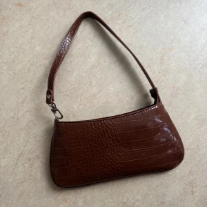 Brun axelväska i croco-imitation - Snygg brun axelväska med croco-mönstrad yta och glansig finish. Väskan har en avtagbar axelrem med silverfärgad detalj och en stilren, avlång form som passar perfekt till din outfit.