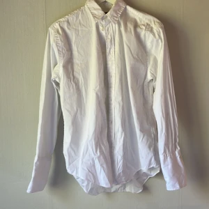 Vit skjorta från Polo Ralph Lauren - Klassisk vit skjorta från Polo Ralph Lauren i bomull. Skjortan har lång ärm, knappar framtill och en snygg krage. Perfekt för en clean och stilren look. Passar dig som gillar tidlös stil och vill ha något som funkar till många outfits.