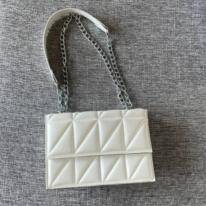Vit quiltad axelväska med kedja - Snygg vit axelväska med quiltat mönster och silverfärgad kedjerem. Väskan har en stilren, rektangulär form och är tillverkad i skinnimitation. Perfekt för dig som vill ha en trendig och clean accessoar till din outfit.