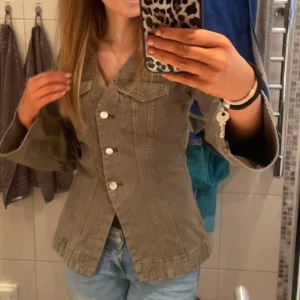  jeansjacka  - Säljer en grön jeansjacka med snygga, vida ärmar och markerad midja. Jackan har stora vita knappar framtill och klassiska bröstfickor. Perfekt för dig som vill ha en unik twist på den klassiska jeansjackan.