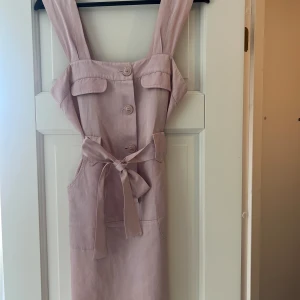 Ljusrosa kort klänning med knytband - Supersöt ljusrosa klänning med breda axelband, knappar framtill och knytband i midjan. Klänningen har fickor och dekorativa bröstfickor. Perfekt för varma dagar när du vill känna dig både chill och stylish.