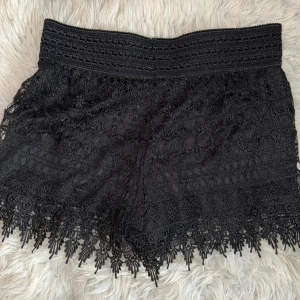 Svarta spets-shorts från Hunkemöller - Svarta shorts från Hunkemöller med bred resår i midjan och detaljerad spets över hela plagget. Kanten nertill har snygga fransar och mönstrad spets som ger en extra touch. Perfekta för dig som vill ha något både bekvämt och stilrent.