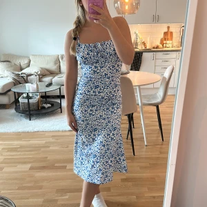 Blommig vit och blå midiklänning - Supersöt vit klänning med blått blommönster och smala axelband. Klänningen har en figurnära passform, knytning vid bysten och är ärmlös!