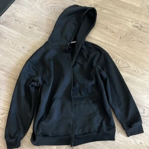 Svart hoodie med dragkedja - Klassisk svart hoodie med dragkedja och huva. Tröjan har två stora fickor framtill och ribbade muddar vid ärmslut och nederkant. Perfekt för en chill och avslappnad stil.