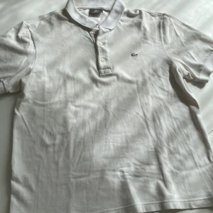 Vit pikétröja från Lacoste - Klassisk vit pikétröja från Lacoste med korta ärmar och broderad krokodil-logga på bröstet. Tröjan har krage, tre knappar framtill och är gjord i mjuk bomull. Perfekt för en clean och stilren look.