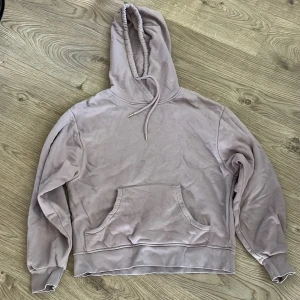 Ljuslila hoodie med magficka - Säljer en ljuslila hoodie med huva och snörning. Tröjan har en stor magficka framtill och ribbade muddar vid ärmslut och nederkant. Perfekt för en chill och avslappnad stil.