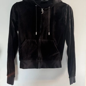 Svart velour hoodie Juicy Couture XS - Svart hoodie från Juicy Couture i mjuk velour med dragkedja, huva och snörning. Klassisk känguruficka framtill och diskret JC-broderi på bröstet. Perfekt för en chill och trendig look.