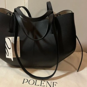 Polène väska - Snygg svart handväska från Polène med stilren design och rundade former. Väskan är tillverkad i strukturerat skinn och har både korta och långa handtag för olika bärsätt. Perfekt för dig som gillar minimalistisk och lyxig look.