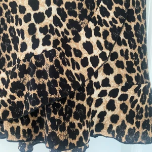 Leopardmönstrad kortkjol med volang - Snygg kortkjol med leopardmönster i svart och beige. Kjolen har volangdetaljer nertill och omlottkänsla framtill. Perfekt för dig som vill sticka ut och addera lite edge till din outfit.