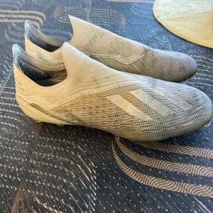 Vita adidas fotbollsskor utan snörning - Säljer ett par vita adidas fotbollsskor med stilren design och diskreta ränder på sidan. Skorna har en strumpliknande passform utan snörning och är tillverkade i syntetmaterial med mönstrad ovandel. Perfekta för dig som vill ha en clean look på planen.