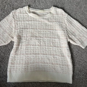 Vit stickad kortärmad tröja - Säljer en vit stickad tröja med korta ärmar och ribbade muddar. Tröjan har ett rutigt mönster och inslag av glittriga trådar som ger en snygg effekt. Perfekt för dig som gillar en stilren men ändå unik look.