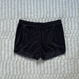 Svarta sammets-shorts med spets - Mysiga svarta shorts i sammetsmaterial från Isabell, storlek M. De har snörning i midjan och fina spetsdetaljer längs kanterna för en extra touch. Perfekta för chill dagar eller när du vill känna dig extra comfy hemma.