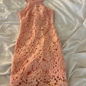 Rosa spetsklänning med blommönster - Supersöt kort rosa klänning med blommig spets och ärmlös design. Klänningen har hög hals och figurnära passform, perfekt för dig som vill sticka ut. Detaljerad spets ger en lyxig känsla och den ljusa färgen är riktigt fräsch till sommaren.