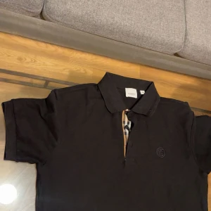 Svart pikétröja från Burberry - Snygg svart pikétröja från Burberry med klassisk krage och korta ärmar. Tröjan har Burberrys ikoniska rutiga detalj vid knäppningen och en diskret broderad logga på bröstet. Perfekt för en clean och stilren look.